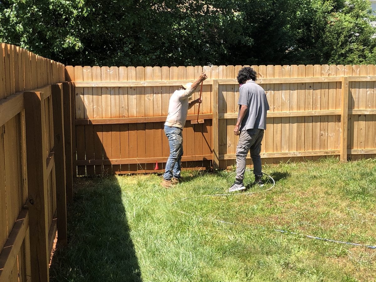 Two painters staining a board fence panel by panel